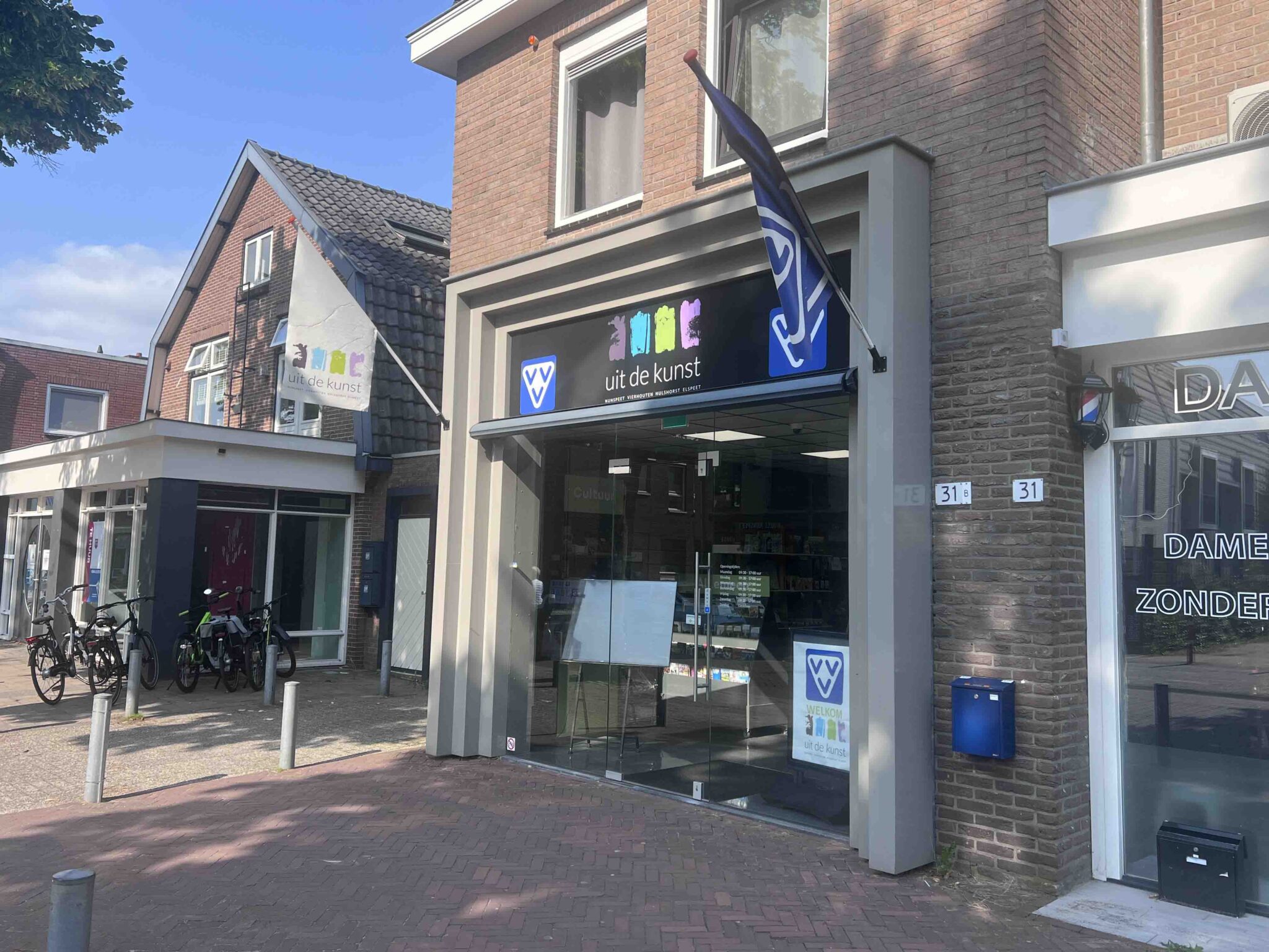 Voorzijde van het pand van VVV Nunspeet uit de Kunst aan de Stationslaan, met vlag en logo zichtbaar naast de ingang